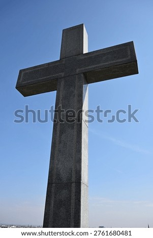 Low angle view of a large gray stone cross standing tall against a clear blue sky. The monumental religious structure features a simple geometric design with visible stone texture and subtle cityscape in the distant background.