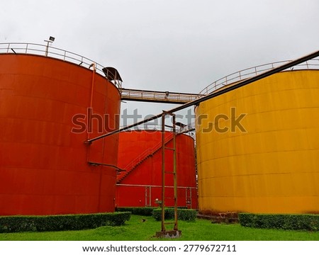 Low angle view of large red, orange, and yellow industrial storage tanks for crude palm oil or CPO at a factory. Metal silos with pipelines, stairs, and walkways under overcast sky. Palm oil industry and agricultural commodity infrastructure.