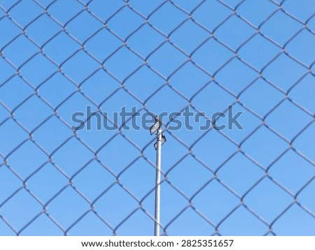 Low angle view of LED floodlight pole framed through blurred chain link fence against blue sky for security concept.