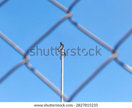 Low angle view of LED floodlight pole framed through blurred chain link fence against clear blue sky for security concept.