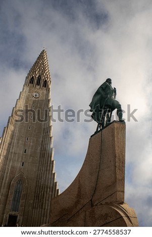 Low angle view of the Leif Erikson statue in front of Hallgrímskirkja church in Reykjavik, Iceland. The statue stands prominently against a partly cloudy sky while the iconic church tower rises in the background. A striking composition highlighting Icelandic history, exploration, and modern architecture, ideal for travel and cultural themes.