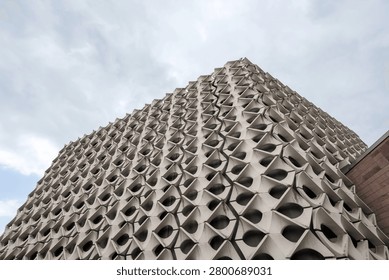 Low angle view looking up at the corner of a brutalist concrete building structure showcasing a complex decorative three-dimensional geometric facade under a cloudy sky.