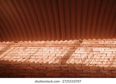 A low angle view looking up at a corrugated roof overhang above a textured red brick wall, where bright sunlight casts distinct linear shadows across the rough masonry surface, highlighting the warm clay tones.