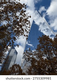 Low angle view looking up at One World Trade Center (Freedom Tower) in New York City. The glass skyscraper reflects a cloudy blue sky and is framed by colorful autumn leaves and neighboring buildings.