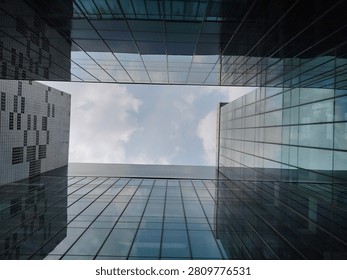 A low angle view looking up towards the facade of modern glass office buildings, reflecting the cloudy blue sky between the structures.
