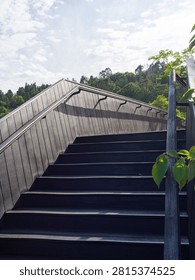 Low angle view looking up wide dark wooden stairs with metal handrails leading toward hilltop under bright cloudy sky. Represents upward mobility, outdoor pedestrian infrastructure, landscape design, and progress concepts.