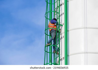 Low angle view of maintenance worker with safety equipment is repairing metal stairs on concrete storage silo against blue sky background