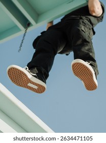 Low angle view of a man hanging from a teal lifeguard tower, showing the soles of his sneakers against a clear blue sky. Casual streetwear moment with urban energy and bold perspective.