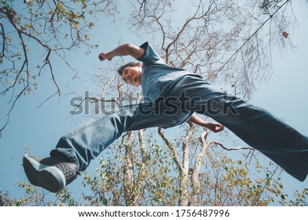 Low angle view of man midair by jumping, crossing step over the camera shot below in forest with tree and sky overhead in concept travel, active lifestyle, overcome obstacles in life or future