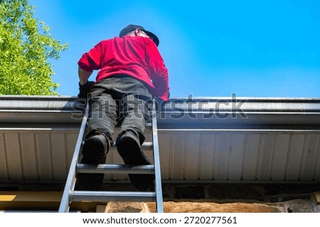 Low angle view of man on ladder cleaning gutters of old stone home with large leafy branches overhead, blue sky and clouds. Architectural features soffit and roof