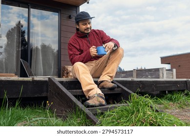 Low Angle View of Man Sitting on Cabin Steps with Laptop and Looking Down at a Blue Coffee Mug