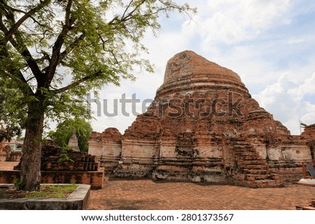 Low angle view of a massive ancient ruined brick stupa at Wat Kudi Dao in Ayutthaya, Thailand. A large green tree stands beside the historic structure on a brick-paved courtyard under cloudy blue sky