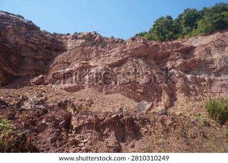 Low angle view of a massive reddish brown sedimentary rock cliff showing stratified layers and weathered rocky mountain terrain under a clear blue sky.