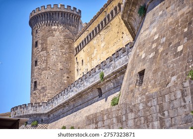 Low angle view of a medieval stone fortress featuring a prominent cylindrical tower with crenellated battlements at the top. The ancient structure displays weathered stone walls in varying shades of brown, beige and gray, with architectural details including arched windows along the upper gallery, defensive ramparts with merlons, and fortified walkways. The tower features narrow vertical windows and a distinctive rounded top. Adjacent walls show signs of age with patches of discoloration, exposed masonry, and small patches of vegetation growing in crevices. A small window with metal bars is visible in the foreground wall. Clear blue sky background emphasizes the imposing height and historic military architecture. The construction exhibits traditional medieval fortification techniques with thick stone walls designed for defense.