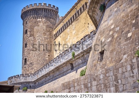 Low angle view of a medieval stone fortress featuring a prominent cylindrical tower with crenellated battlements at the top. The ancient structure displays weathered stone walls in varying shades of brown, beige and gray, with architectural details including arched windows along the upper gallery, defensive ramparts with merlons, and fortified walkways. The tower features narrow vertical windows and a distinctive rounded top. Adjacent walls show signs of age with patches of discoloration, exposed masonry, and small patches of vegetation growing in crevices. A small window with metal bars is visible in the foreground wall. Clear blue sky background emphasizes the imposing height and historic military architecture. The construction exhibits traditional medieval fortification techniques with thick stone walls designed for defense.