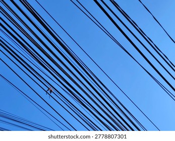 Low angle view of messy electrical power lines and telecommunication cables crisscrossing against a clear blue sky. The tangled black wires create a complex geometric pattern and abstract urban industrial background