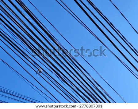 Low angle view of messy electrical power lines and telecommunication cables crisscrossing against a clear blue sky. The tangled black wires create a complex geometric pattern and abstract urban industrial background