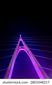 Low angle view of a modern cable-stayed bridge tower illuminated with vibrant purple lights against a dark night sky, showcasing contemporary urban architecture and engineering design.