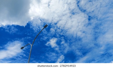 Low angle view of modern double street light pole against bright blue sky with scattered white clouds. Urban infrastructure and outdoor lighting concept with clear weather and open sky background