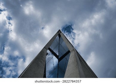 Low angle view of a modern triangular concrete and glass architectural structure pointing towards a dramatic cloudy sky.