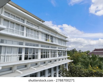 Low angle view of a modern white multi-story academic or office building facade under a bright blue sky with tropical green trees
