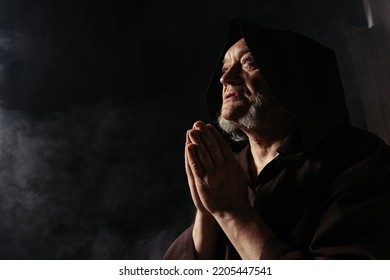 Low Angle View Of Monk In Black Hooded Robe Praying On Dark Background