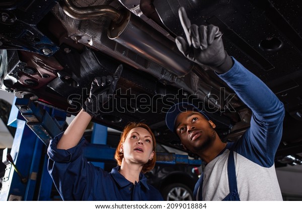Low angle view of multiethnic\
mechanics in gloves pointing with fingers under car in\
service