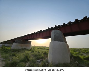 Low angle view of an old iron girder railway bridge supported by concrete pillars crossing over a green rural landscape under a clear blue sky.