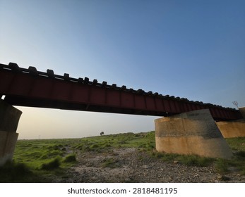 Low angle view of an old iron girder railway bridge supported by concrete pillars crossing over a green rural landscape under a clear blue sky.