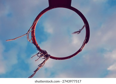 Low angle view of an old red basketball hoop with a torn net against a blue cloudy sky