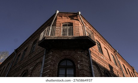 Low angle view of an old red brick building corner with a vintage balcony against a deep blue sky.