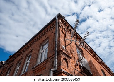 Low angle view of an old red brick building corner with windows, drainpipe and rooftop antennas against a cloudy blue sky.