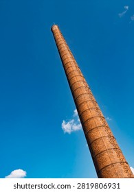 Low angle view of an old red brick industrial chimney reaching into a bright blue sky. Historic smokestack architecture with climbing ladder.