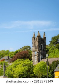 Low Angle View Of Old Stone Church Bell Tower With Spires And Clock Face Surrounded By Green Trees Under Bright Blue Sky With Wispy Clouds