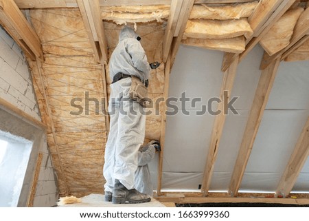 Low angle view of one foreman in overalls working with rockwool insulation material, standing inside new house under construction, fasten warmth layer material on roof ceiling and wall