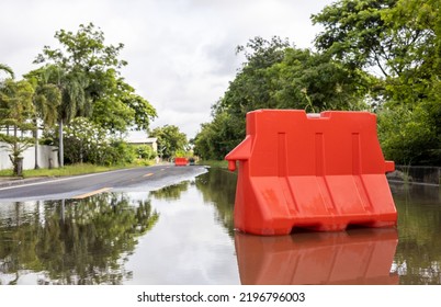 A low angle view of orange plastic barricades placed on a flooded road with water reflections near a tree to prevent vehicles from passing through a rural area in Thailand.