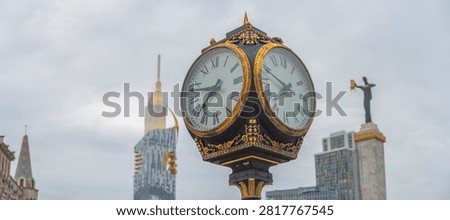 Low angle view of an ornate black and gold retro street clock at Europe Square in Batumi, Georgia, with blurred city landmarks under a cloudy sky