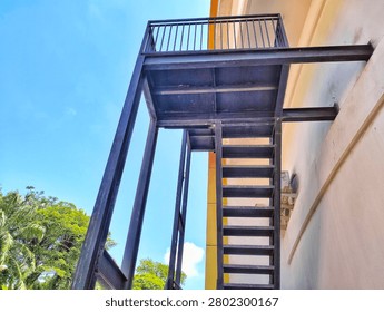Low angle view of an outdoor black metal emergency fire escape staircase attached to a building wall against a clear blue sky.
