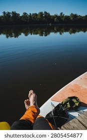 Low angle view from paddleboard shows relaxed barefoot feet and orange pants resting at board edge, calm dark lake reflecting treeline and blue sky, with dock gear, shoes and wood planks nearby
