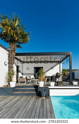 Low angle view of a palm tree rising against a black aluminum bioclimatic pergola, with a wooden deck, potted ponytail palms and outdoor lounge area in a contemporary residential garden under a clear blue sky.