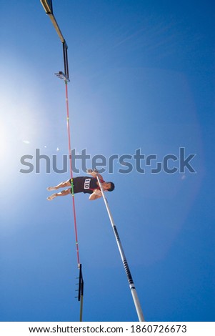 Low angle view of a pole vault athlete going over bar with a clear blue sky and lens flare in the background.
