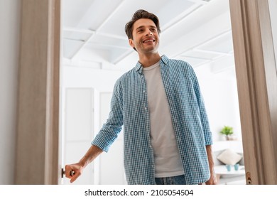 Low Angle View Portrait Of Cheerful Casual Man Receiving Visitor, Happy Young Guy Standing In Doorway Of Modern Apartment, Millennial Male Holding Door Looking Out Of His Flat