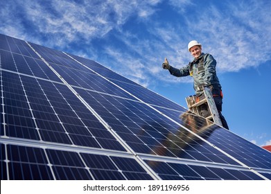 Low angle view portrait of a smiling worker, installing solar batteries, who is standing on ladder at solar plant against blue sky, showing thumb up. Concept of alternative sources of energy.