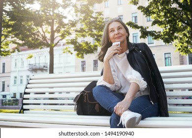 Low Angle View Of Positive Stylish Middle Aged Female In Trendy Leather Jacket, Blue Jeans And Sneakers Having Pensive Look, Sitting Comfortably On Bench With Cell Phone, Thinking Where To Have Lunch