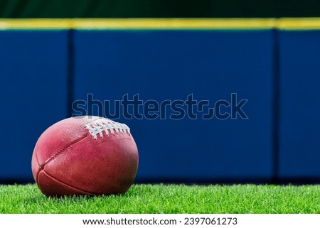 A low angle view of a pro American Football sitting on the turf of a stadium with blue padded wall in the background.