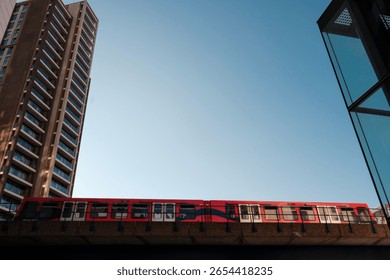 Low angle view of a red and white dlr train moving on an elevated track, passing between modern glass building and residential skyscraper against clear blue sky in london, uk