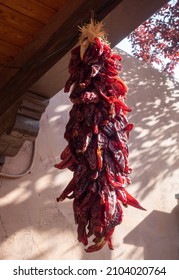 Low angle view of a ristra with drying red chile peppers hanging against a light pink stucco wall with a tree with red leaves in the background.