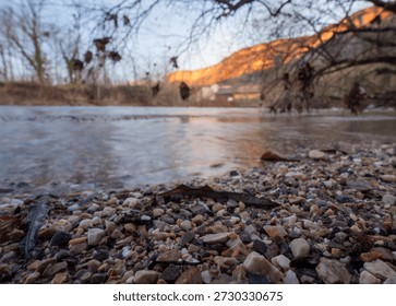 A low angle view of river pebbles in the foreground with a blurred mountain village and glowing orange cliffs at sunset over clear stream water.