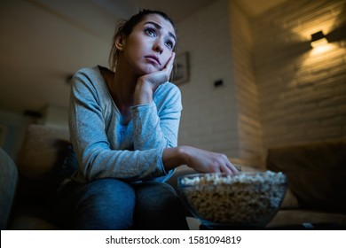 Low angle view of sad woman watching TV and eating popcorn in the evening at home. 