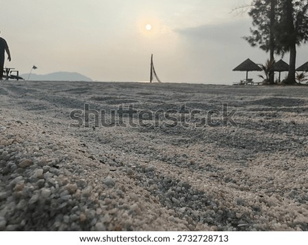 Low angle view of sandy beach at sunrise with soft golden sunlight, peaceful coastal atmosphere, and silhouettes of trees and beach huts creating a calm tropical morning scene.